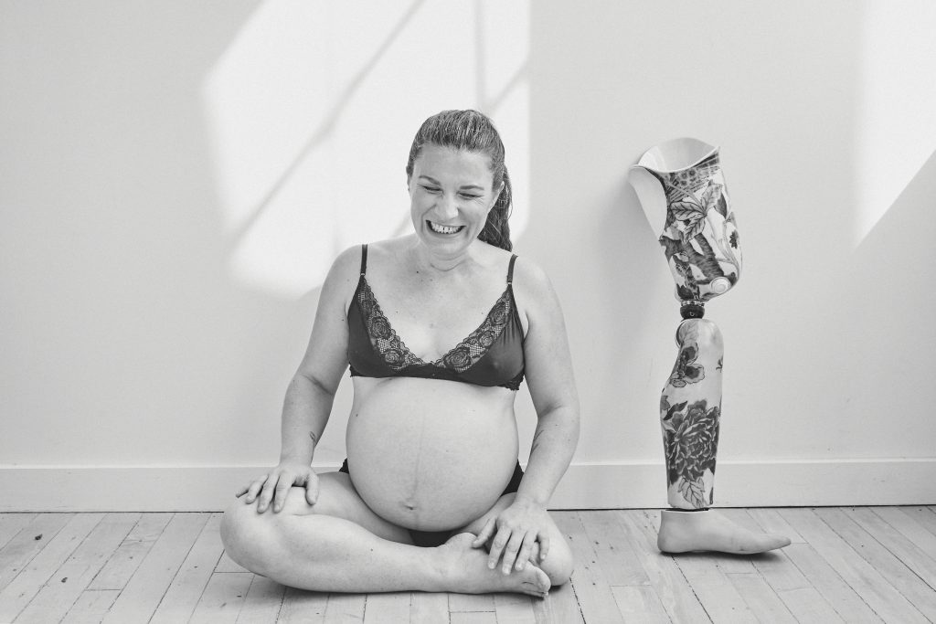 black and white photo of Christa sitting on the ground. Her eyes are closed and she's laughing. Her prosthetic leg is is leaning on the wall behind her.