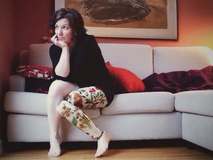 Christa on a couch looking out a window. She wears a black long sleeved top and her legs are bear, showing her floral prosthesis.