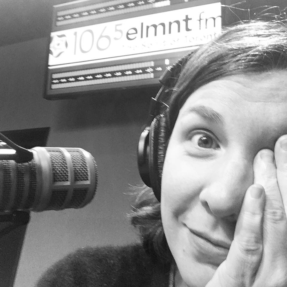 Black and white photo of Christa covering half her face with her hand. A sign in the back says 106.5 ELMNT FM